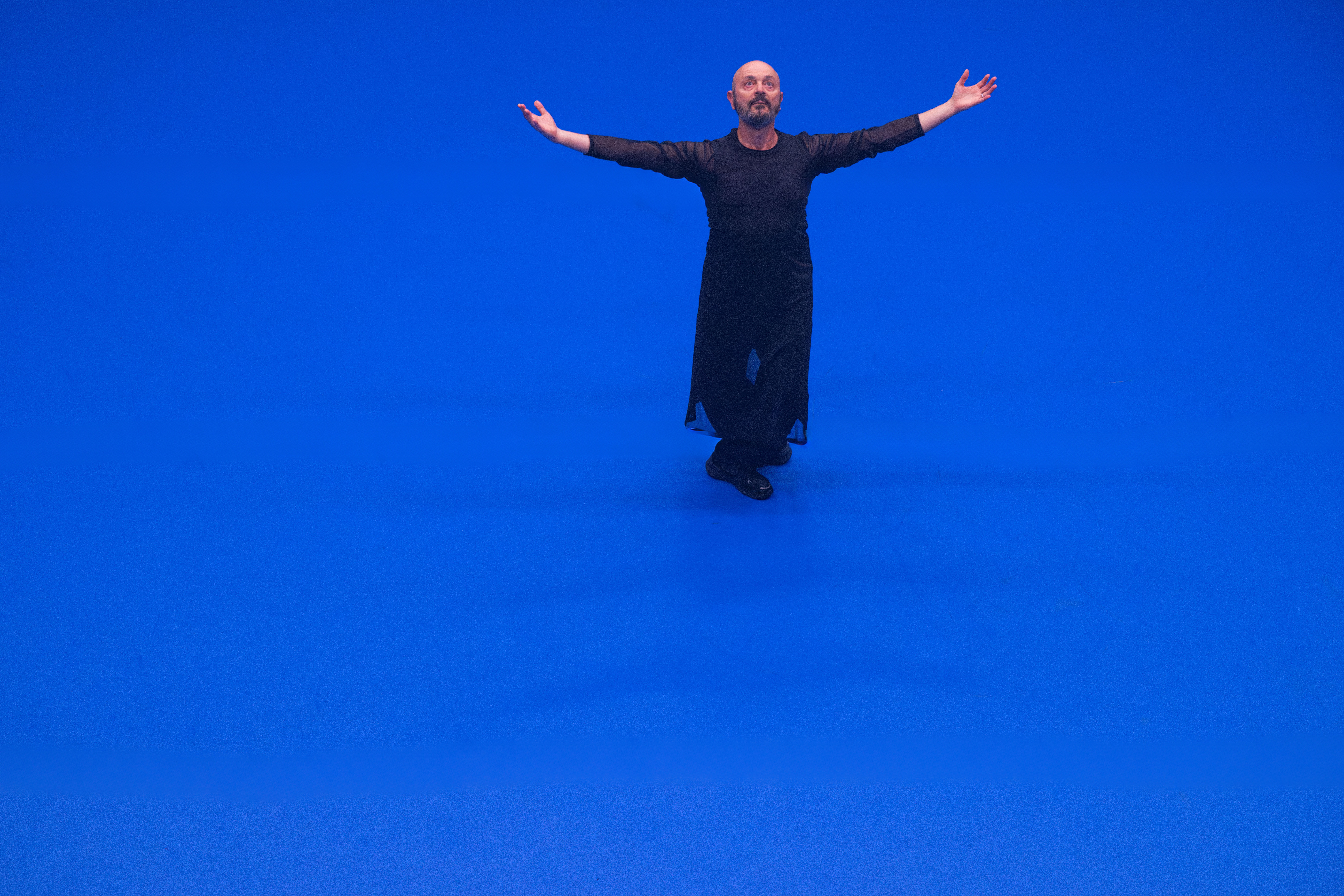 1 performer with black clothes on a bright blue floor, spreading both arms and looking up to the sky